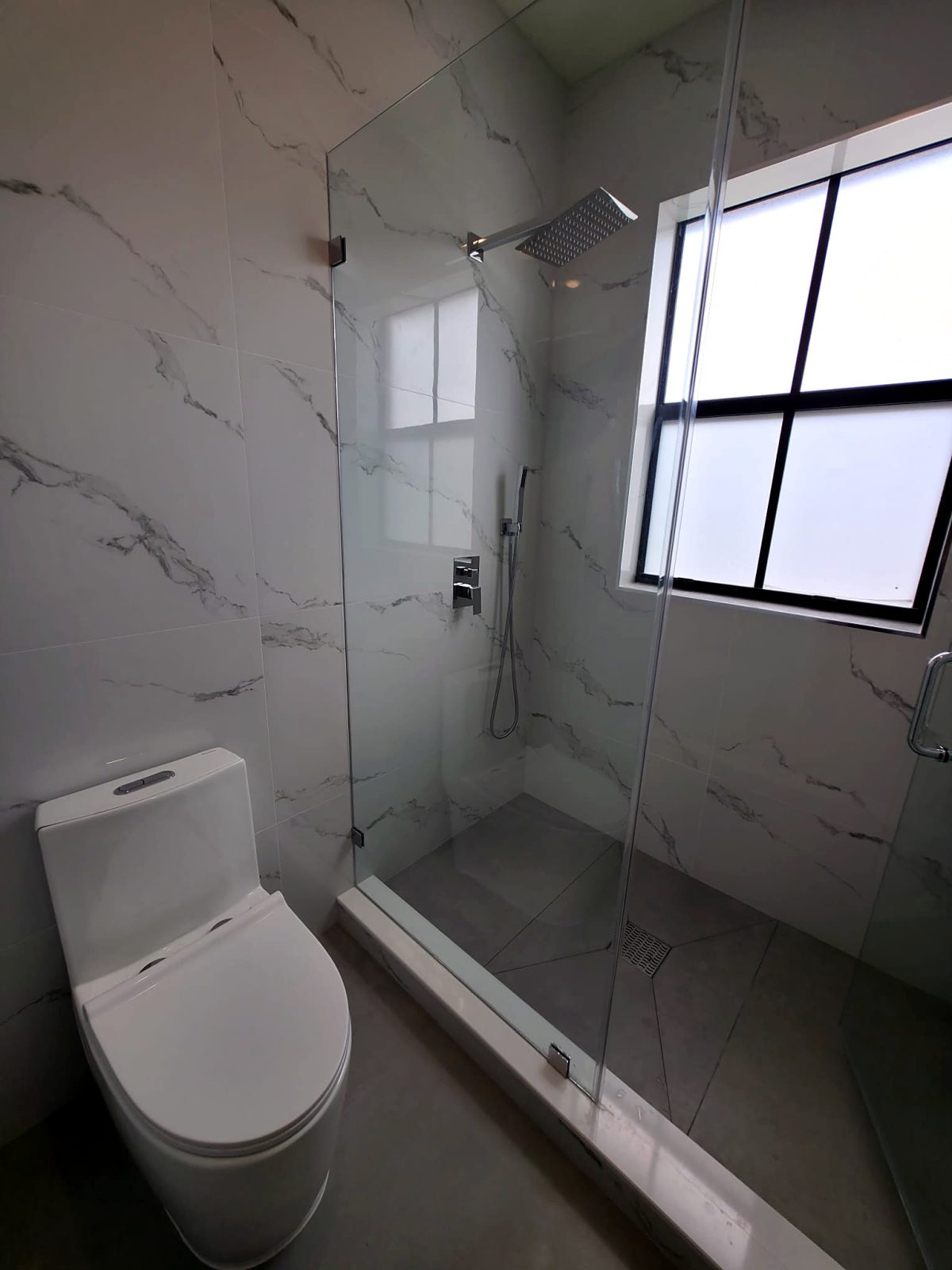 Glass shower next to a one-piece toilet, marble walls